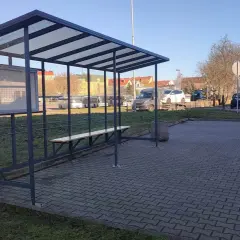 Prosty, tymczasowy dworzec autobusowy z ławką i tablicą ogłoszeń. W tle widać autobusy i samochody.