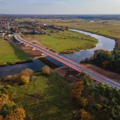 Nowy, czerwony most na warcie przecina spokojną rzekę, łącząc malowniczą wieś z zielonymi polami i lasami jesienią.