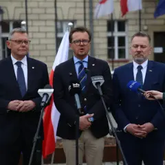 Trzech mężczyzn w garniturach przemawia do mikrofonów. W tle flagi Polski i Unii Europejskiej.