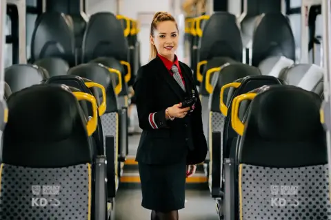 Uśmiechnięta konduktorka w mundurze stoi między rzędami siedzeń w pociągu. Czy u konduktora można płacić blikiem?
