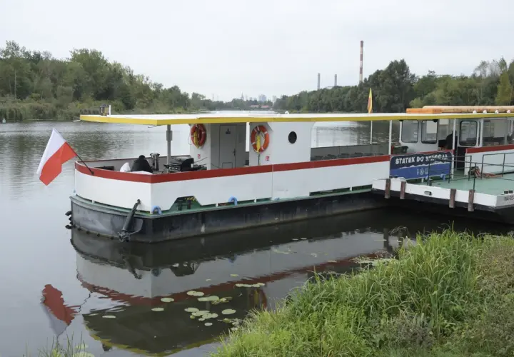 Statek do Serocka z polską flagą czeka na pasażerów. W tle widoczne są kominy elektrowni i drzewa.