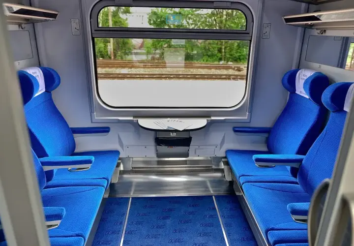 Wnętrze wagonu Intercity z niebieskimi siedzeniami i oknem z widokiem na tory.