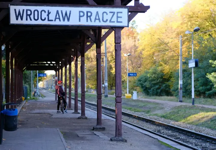 Na stacji Wrocław Pracze, pod drewnianą wiatą, osoba na rowerze czeka na pociąg TLK. Jesienne drzewa w tle.