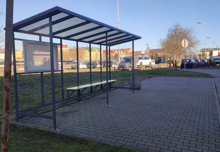Prosty, tymczasowy dworzec autobusowy z ławką i tablicą ogłoszeń. W tle widać autobusy i samochody.