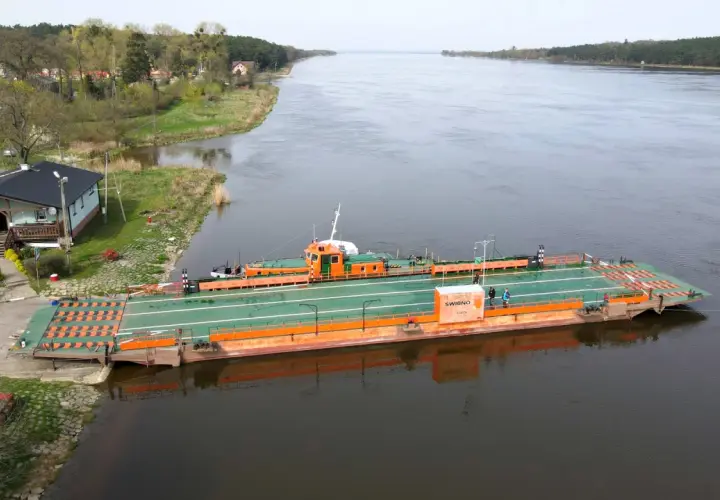 Prom "Świbno" czeka na pasażerów przy brzegu, gotowy do przeprawy przez szeroką rzekę.