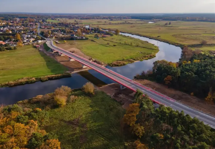 Nowy, czerwony most na warcie przecina spokojną rzekę, łącząc malowniczą wieś z zielonymi polami i lasami jesienią.