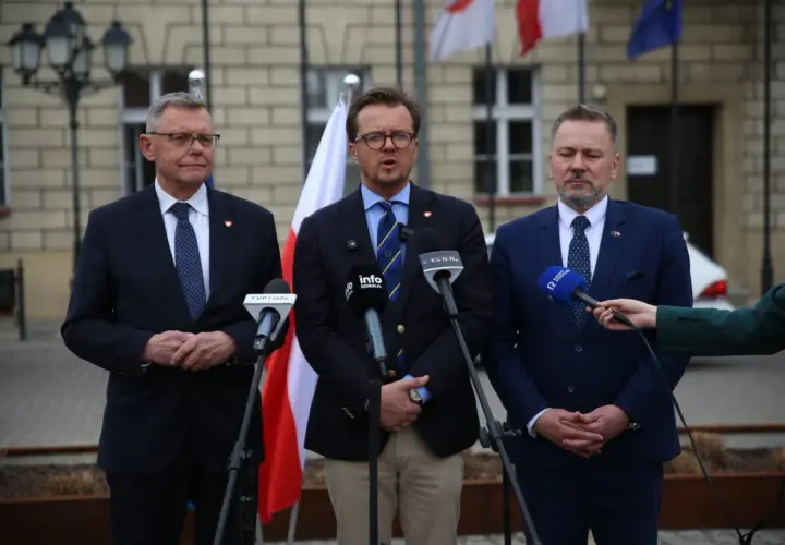 Trzech mężczyzn w garniturach przemawia do mikrofonów. W tle flagi Polski i Unii Europejskiej.