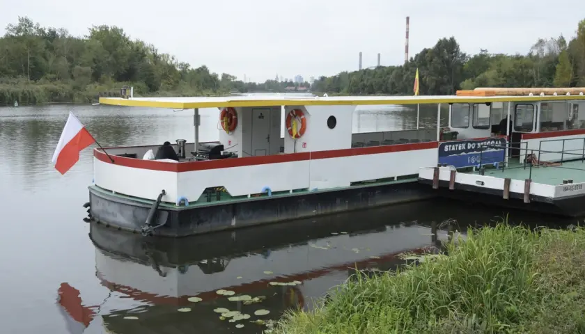Statek do Serocka z polską flagą czeka na pasażerów. W tle widoczne są kominy elektrowni i drzewa.