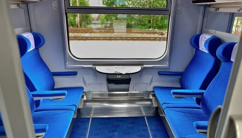 Wnętrze wagonu Intercity z niebieskimi siedzeniami i oknem z widokiem na tory.