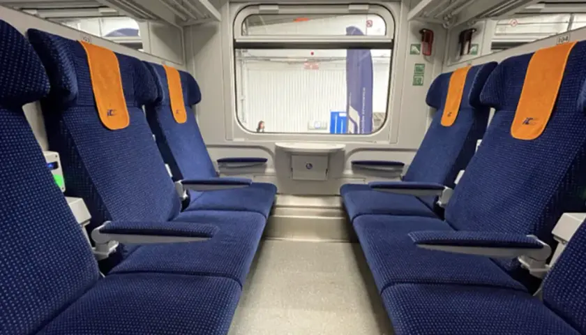 Wnętrze wagonu TLK z niebieskimi siedzeniami i pomarańczowymi zagłówkami. Obok siedzeń widać zielone lampki, co sugeruje, że czy w TLK są kontakty.
