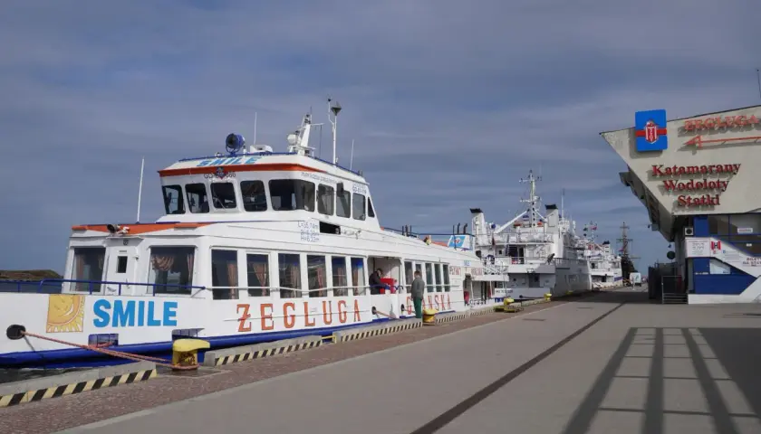 Wodolot Gdynia Hel "SMILE" cumuje przy nabrzeżu. W tle inne statki i budynek z napisem "Katamrany Wodoloty Statki".