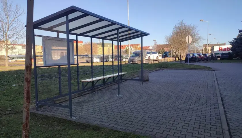 Prosty, tymczasowy dworzec autobusowy z ławką i tablicą ogłoszeń. W tle widać autobusy i samochody.