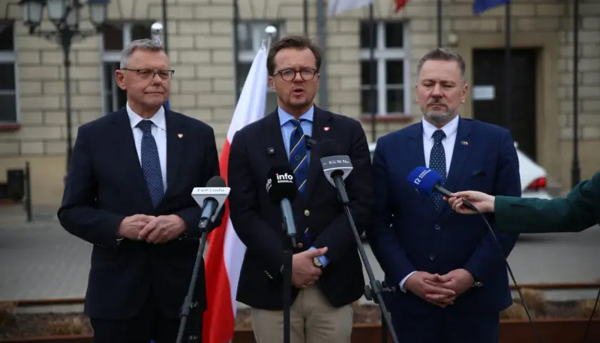 Trzech mężczyzn w garniturach przemawia do mikrofonów. W tle flagi Polski i Unii Europejskiej.