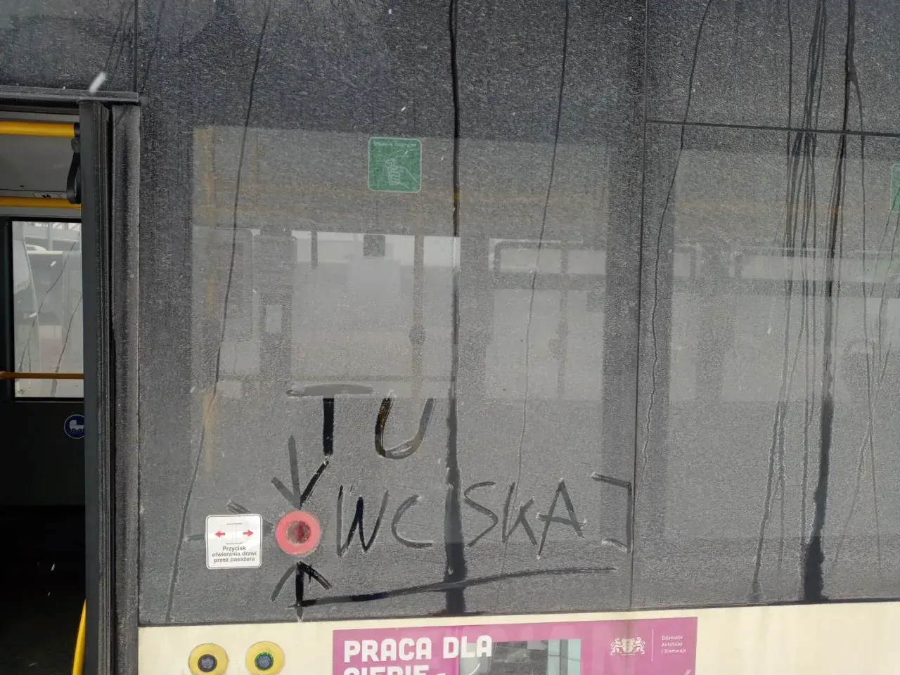Brudne drzwi pociągu z napisem "TU WCISKAJ" i ikoną przycisku otwierania drzwi.