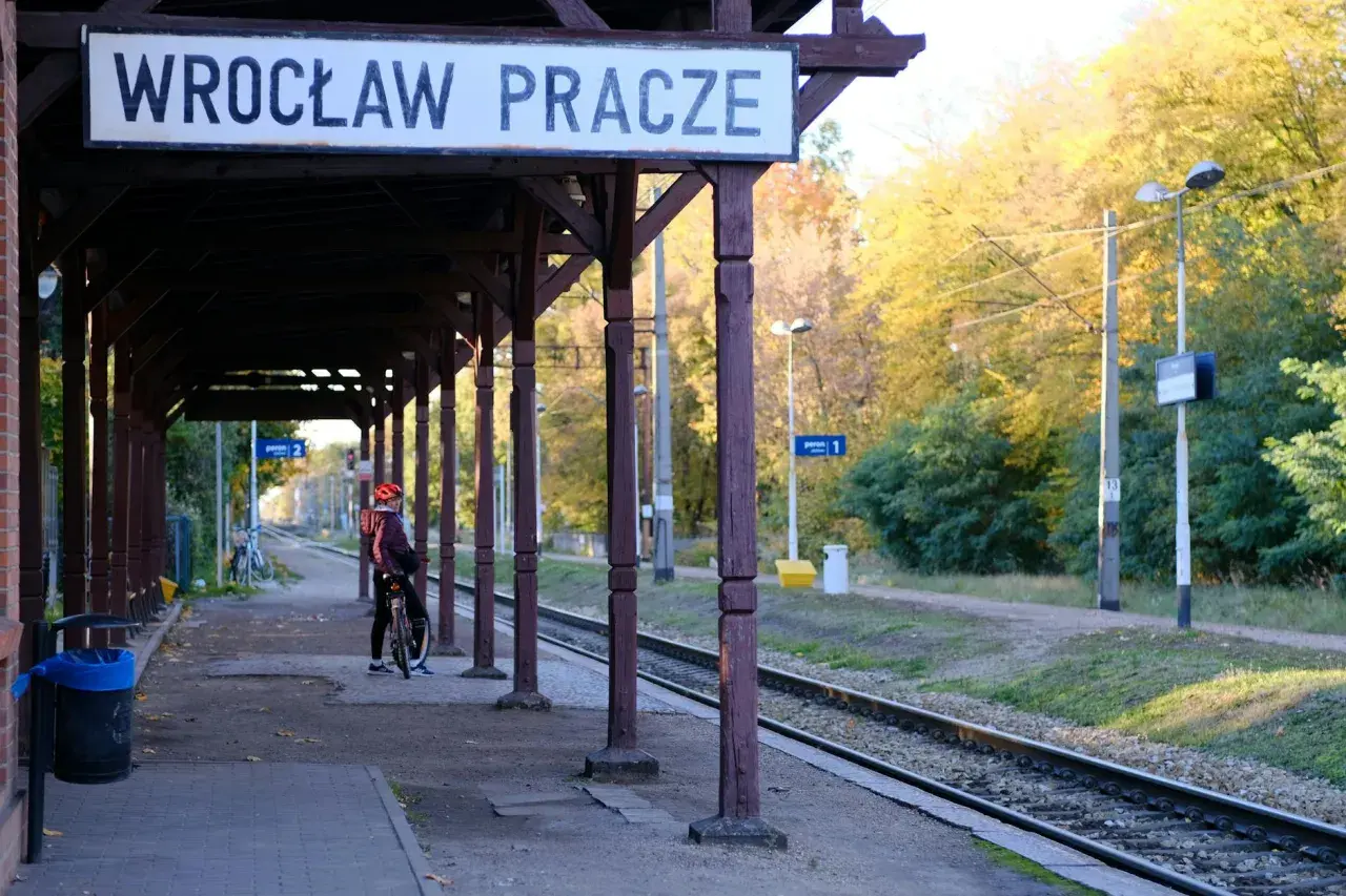 Na stacji Wrocław Pracze, pod drewnianą wiatą, osoba na rowerze czeka na pociąg TLK. Jesienne drzewa w tle.
