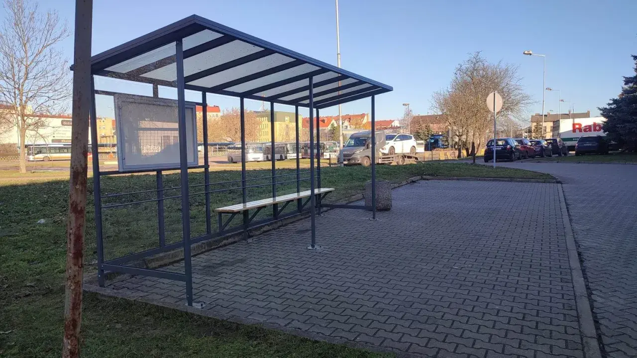 Prosty, tymczasowy dworzec autobusowy z ławką i tablicą ogłoszeń. W tle widać autobusy i samochody.