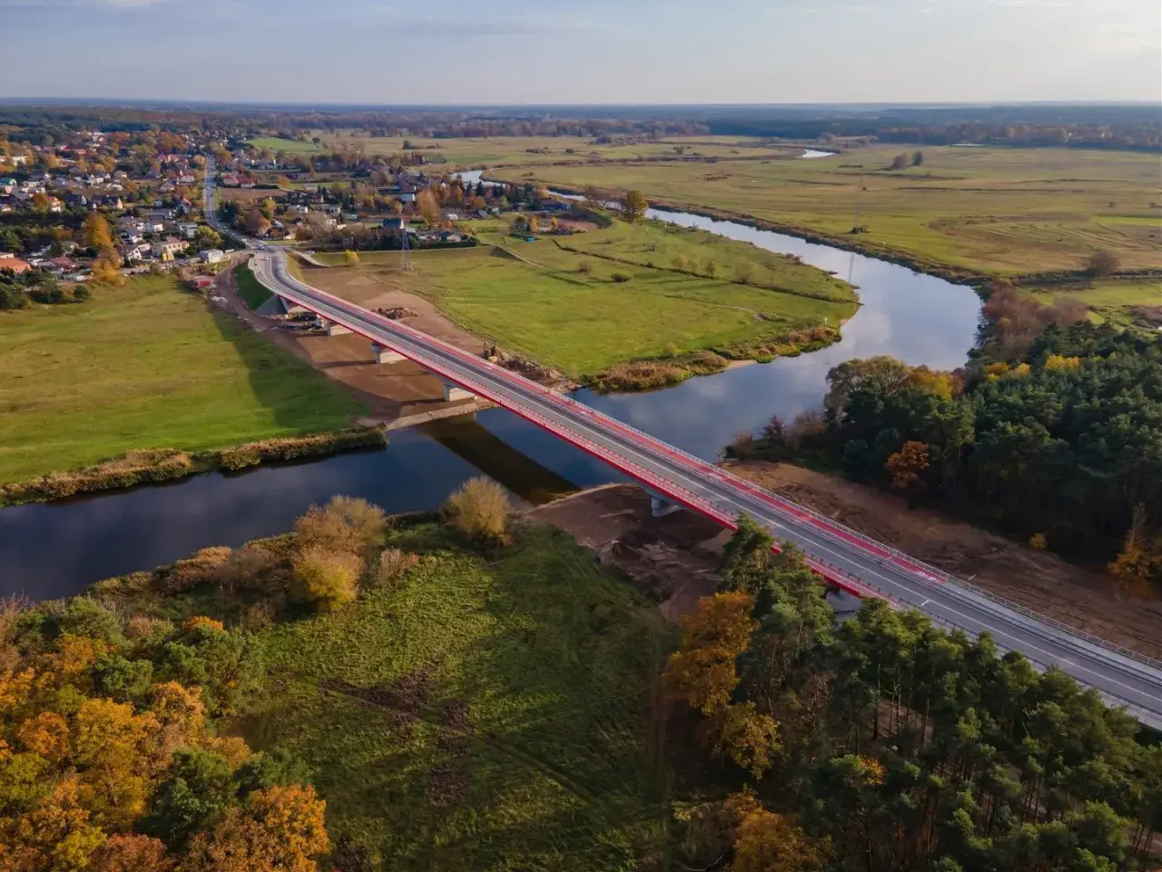 Nowy, czerwony most na warcie przecina spokojną rzekę, łącząc malowniczą wieś z zielonymi polami i lasami jesienią.