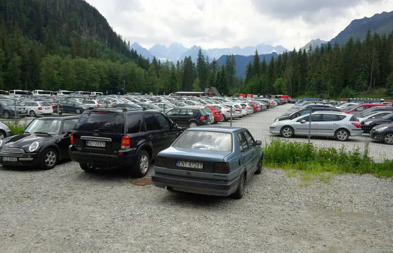 Duży parking samochodowy u podnóża gór, z widokiem na Tatry, jakby eic morskie oko patrzyło z góry.