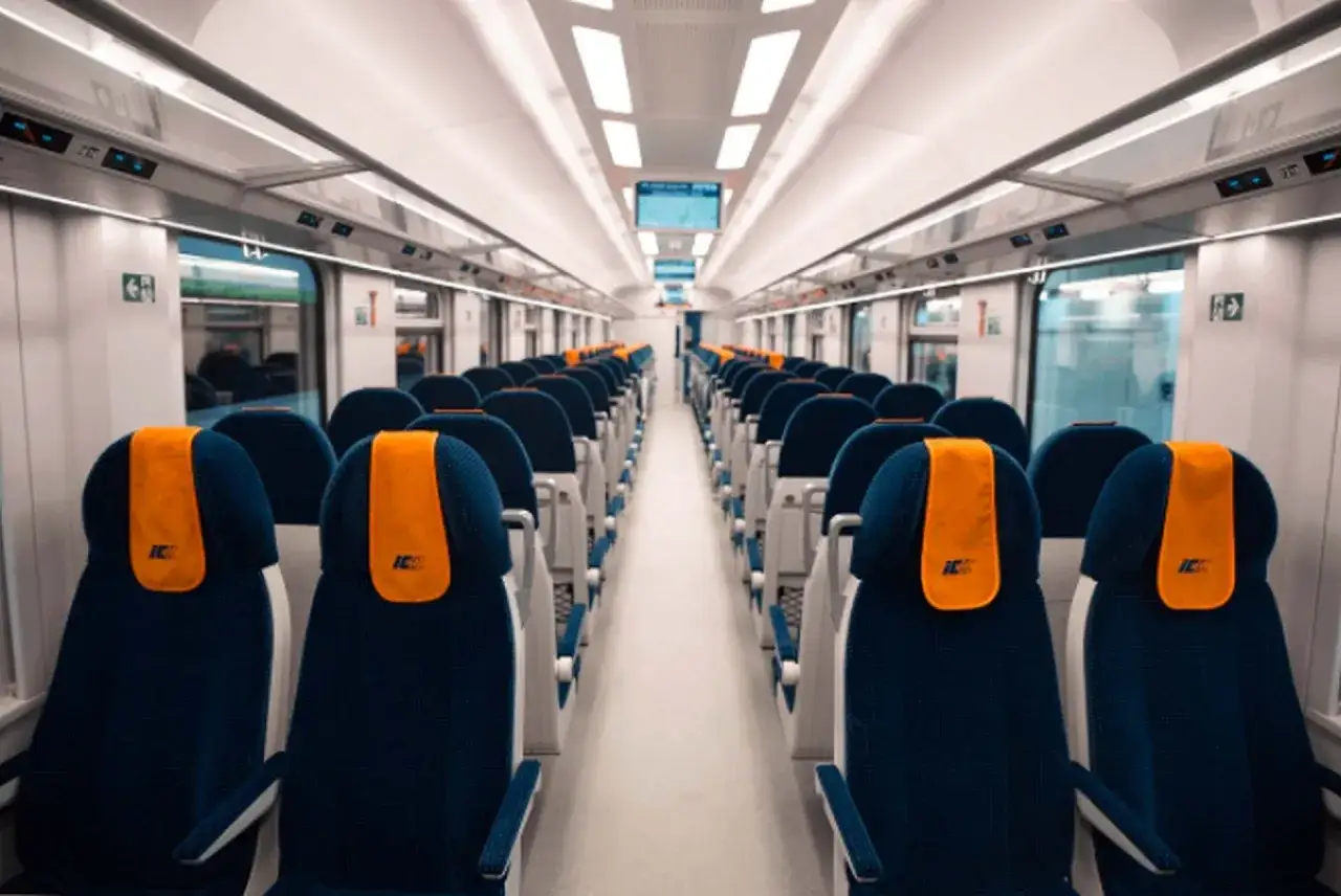 Wnętrze nowoczesnego pociągu Intercity z rzędami granatowych foteli i pomarańczowymi zagłówkami. To właśnie intercity, czyli podróżowanie z klasą.