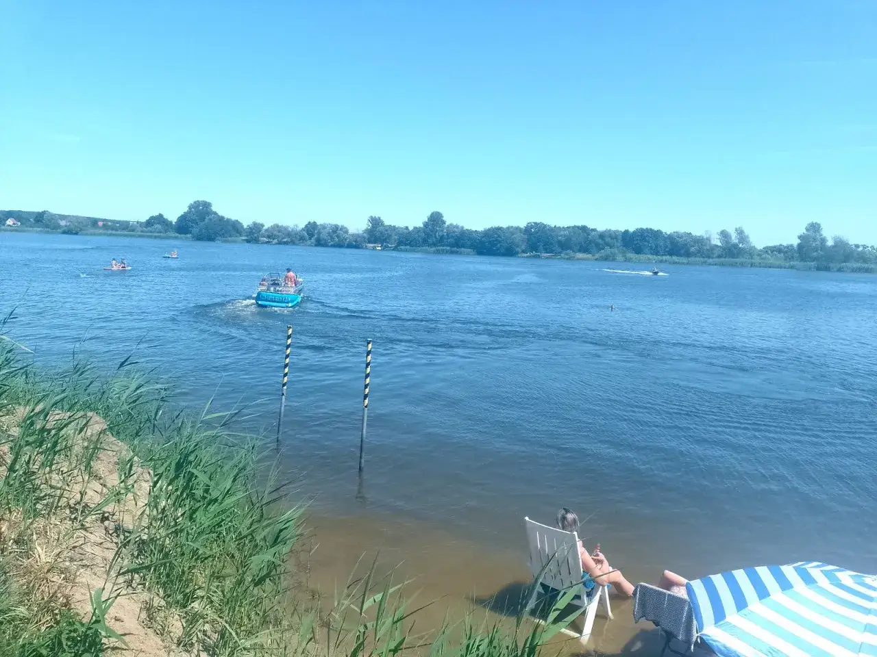 Letnia przeprawa promowa na zalew prężyce. Motorówka z pasażerami płynie po wodzie, na brzegu odpoczywają ludzie.