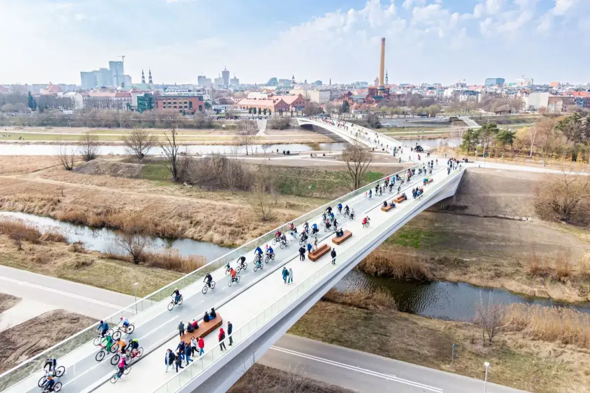 Ludzie na rowerach i piesi korzystają z nowoczesnego mostu na warcie, podziwiając panoramę miasta.