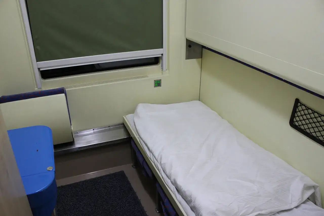 Wnętrze wagonu TLK: łóżko z białym pościelonym posłaniem, niebieska toaleta, okno z zieloną roletą.