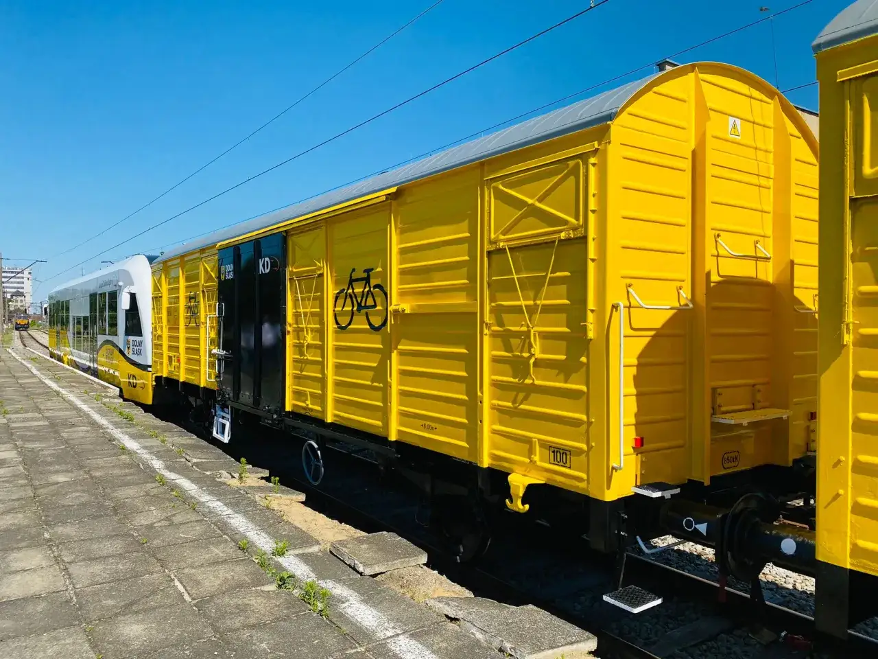 Żółty wagon towarowy z symbolem roweru, część pociągu Koleje Dolnośląskie, stoi na torach przy peronie.