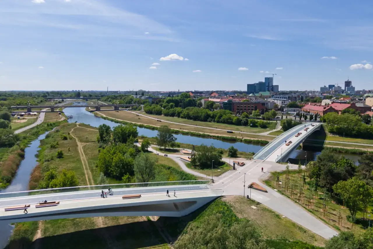 Nowoczesny most na warcie, łączący brzegi rzeki, z widokiem na miasto i zieleń. Ludzie spacerują i jeżdżą na rowerach.