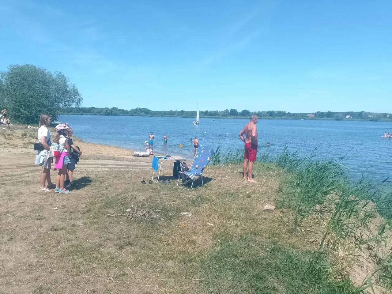 Rodzina na plaży, dzieci bawią się w wodzie, a w tle żaglówka. To idealne miejsce na przeprawę promową na Zalew Prężyce.