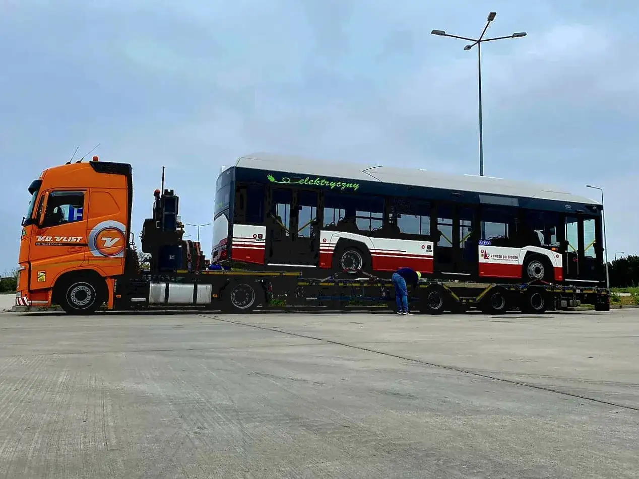 Pomarańczowa ciężarówka transportuje dwa nowe autobusy elektryczne. Jeden z nich jest ładowany na naczepę, a drugi już na niej stoi. W tle widać słup oświetleniowy. To nowy transport dla mzk zajezdnia autobusowa.