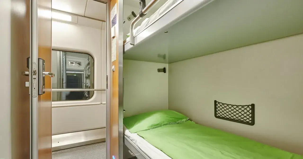 Wnętrze wagonu sypialnego Pendolino z zieloną pościelą i górnym łóżkiem. Drzwi otwarte na korytarz.