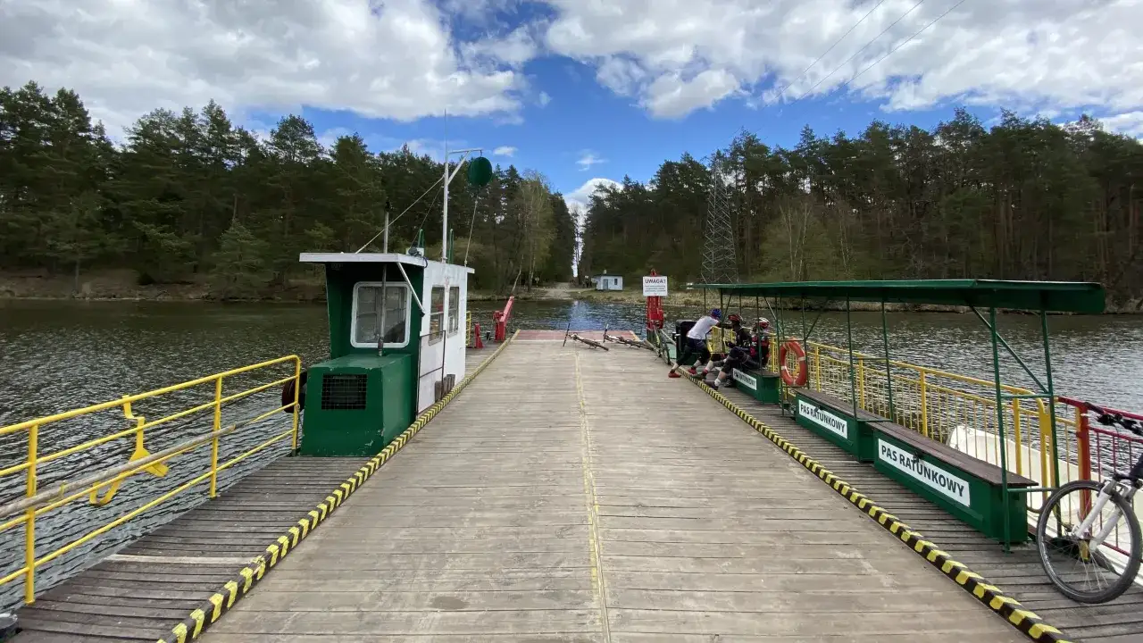 Przeprawa promowa Sokole Kuźnica. Drewniany pomost promu z rowerami i ludźmi czeka na przeprawę przez jezioro.