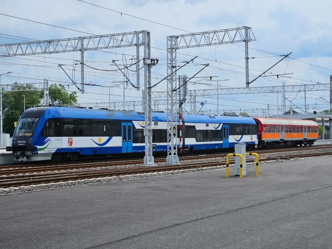 Nowy, niebiesko-biały pociąg stoi na torach, gotowy do podróży. Obok niego widać starszy, pomarańczowo-czerwony wagon. To pociąg do Giżycka.