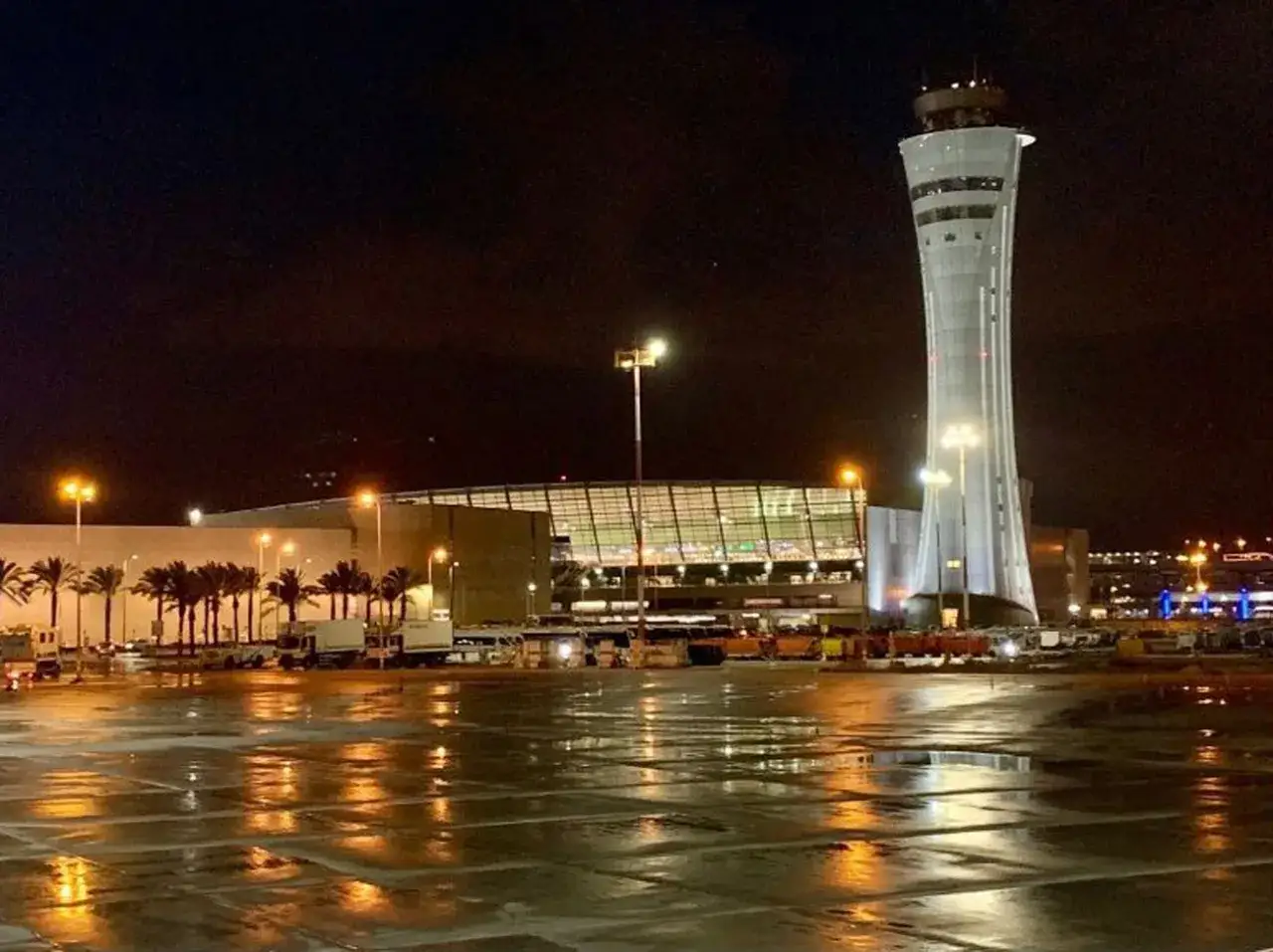 Wieczorem na lotnisku Ben Guriona, wieża kontroli lotów i terminal oświetlone, mokry asfalt odbija światła.