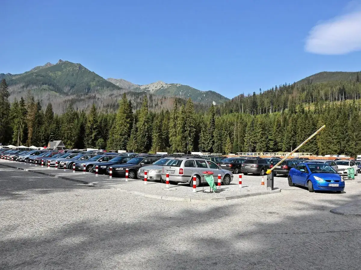 Parking przy Morskim Oku pełen samochodów, w tle góry i las.
