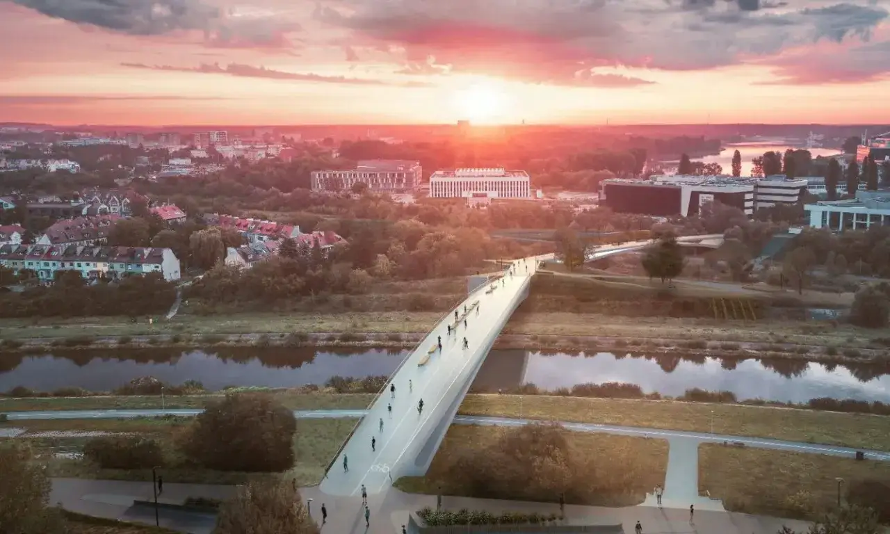 Nowoczesny most na warcie, z ludźmi spacerującymi i jeżdżącymi na rowerach, w otoczeniu zieleni i budynków, podczas zachodu słońca.