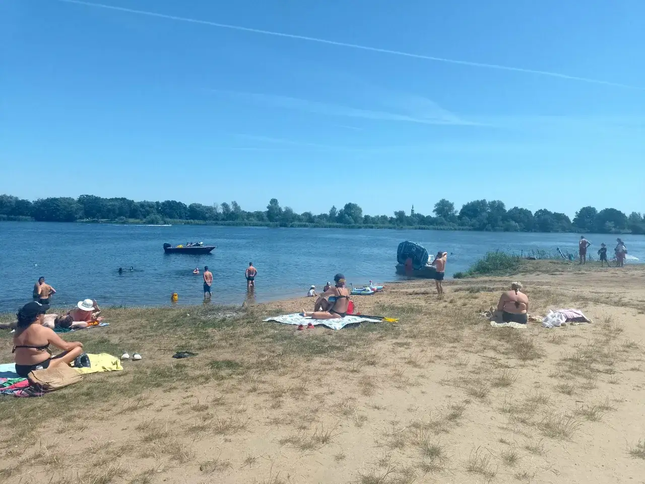 Słoneczny dzień nad zalewem Prężyce. Ludzie odpoczywają na plaży, kąpią się i pływają łódką. Przeprawa promowa na zalew prężyce w tle.