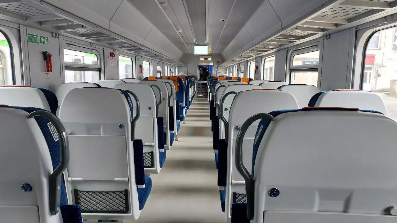 Wnętrze pociągu IC z rzędami białych siedzeń i niebieskimi akcentami. Trudno określić, ile wagonów ma pociąg, ale widać długi korytarz.