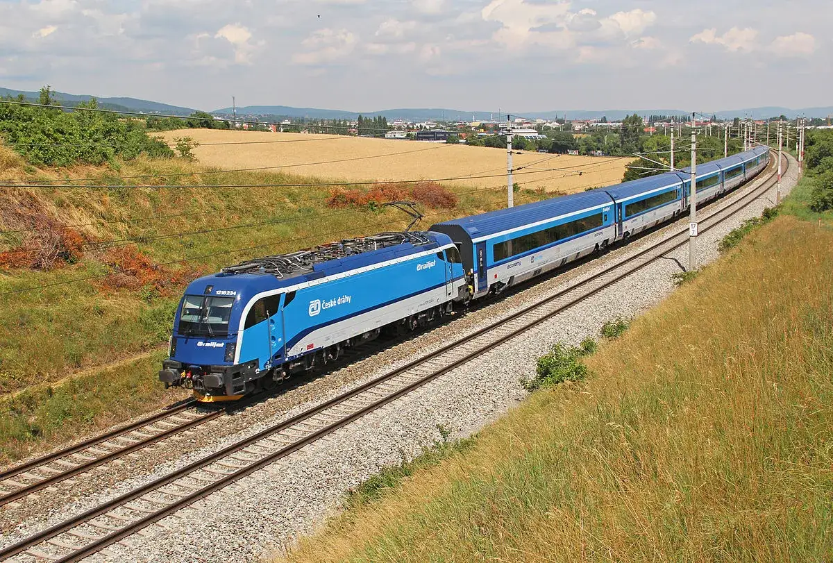 Niebieski pociąg ČD Railjet pędzi po torach, zgodnie z rozkładem jazdy. W tle pola i miasto.