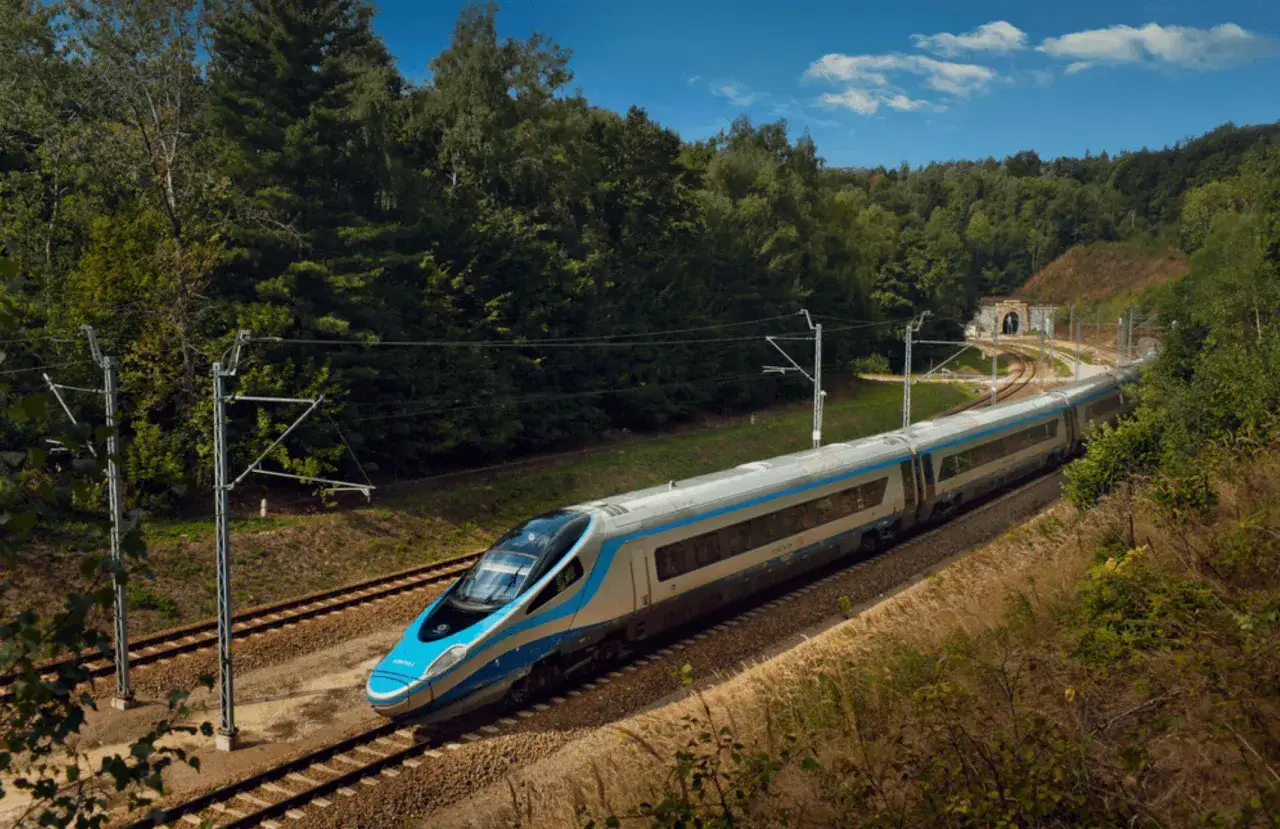 Błękitno-srebrne Pendolino pędzi przez zielony las, zbliżając się do tunelu. Widać, jak szybko jedzie Pendolino.