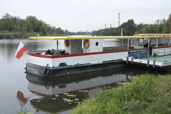Statek do Serocka z polską flagą czeka na pasażerów. W tle widoczne są kominy elektrowni i drzewa.
